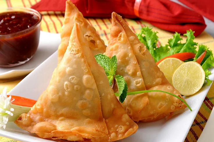 Golden samosas on a white platter with mint, lemon and chutney